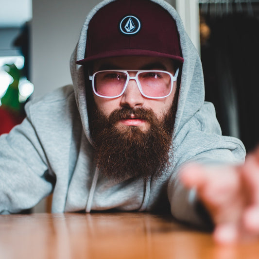 A man with a curly beard and pink lensed sun glasses and maroon baseball cap.